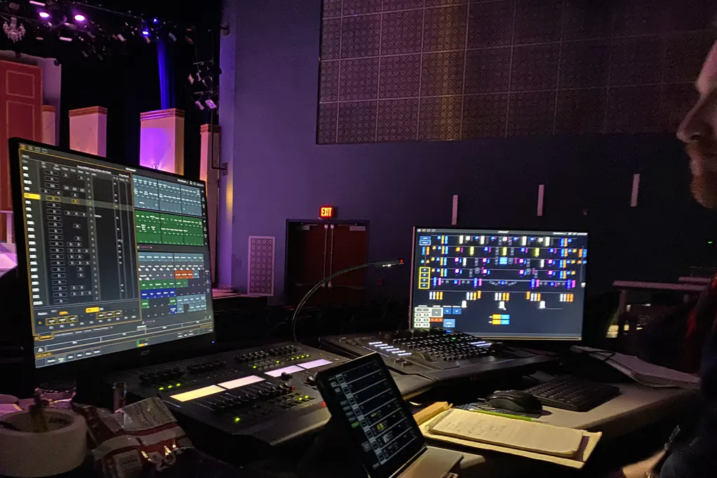 A theatre front-of-house position during a show &mdash; a cue list and a light plot live on two monitors, a console beneath them and a handwritten cue sheet on a clipboard, with the lit set visible beyond.