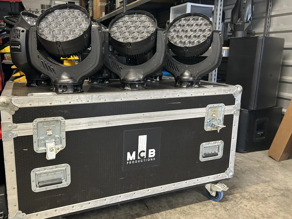 Three LED moving-head wash fixtures with clear multi-lens arrays standing on a wheeled flight case marked with the MCB Productions logo, in front of steel shelving stacked with cased equipment, two loudspeaker cabinets alongside.