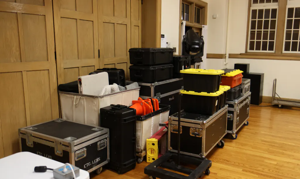 Road cases, wheeled dollies, equipment bins and a moving-head fixture on a stand, staged against the wall of a wood-panelled hall during a load-in.