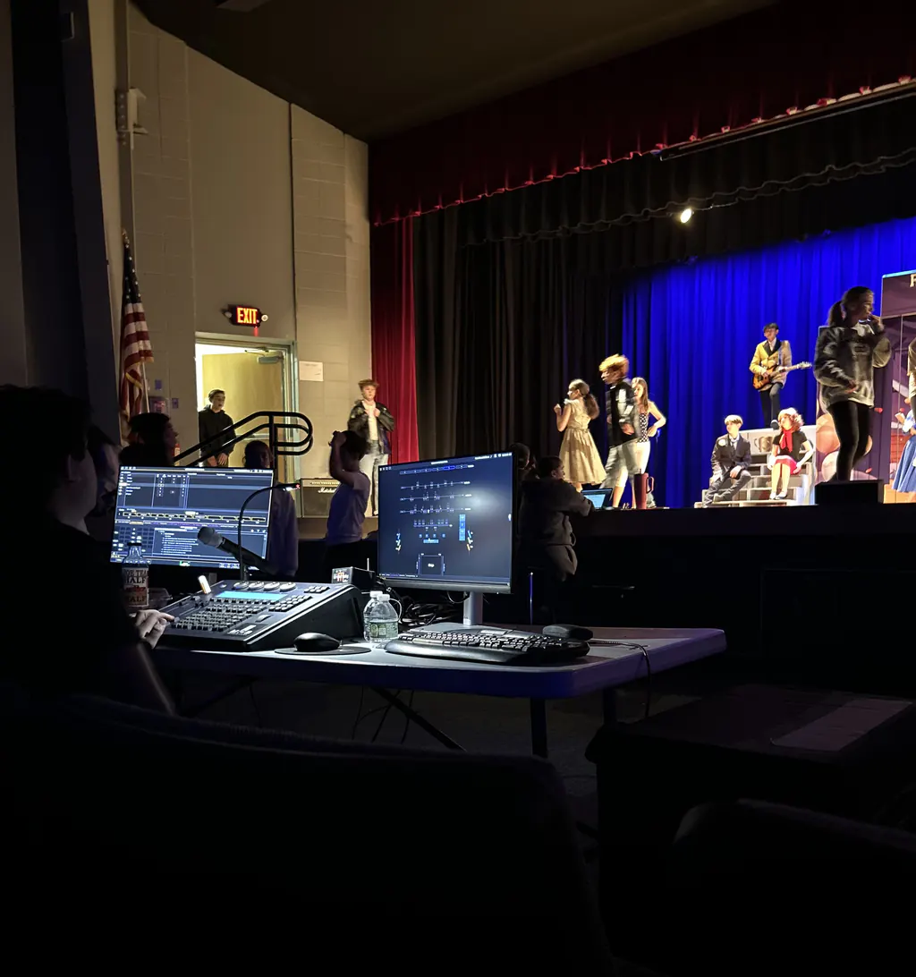 An operator at a lighting desk with a cue list on one monitor and the light plot on the other, calling a show on the lit stage beyond.