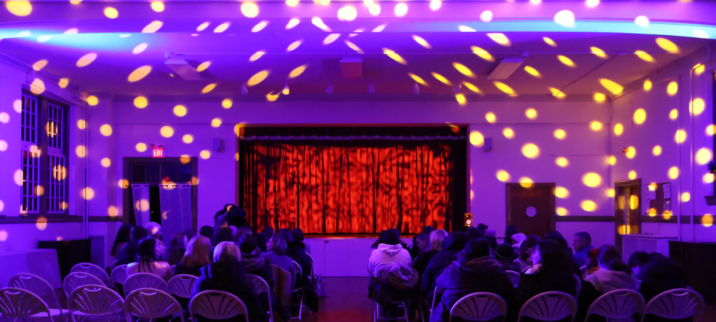 A plain hall of folding chairs with a seated audience, re-lit end to end &mdash; purple wash across every wall, warm break-up patterns thrown across the whole ceiling, and the stage curtain lit red.