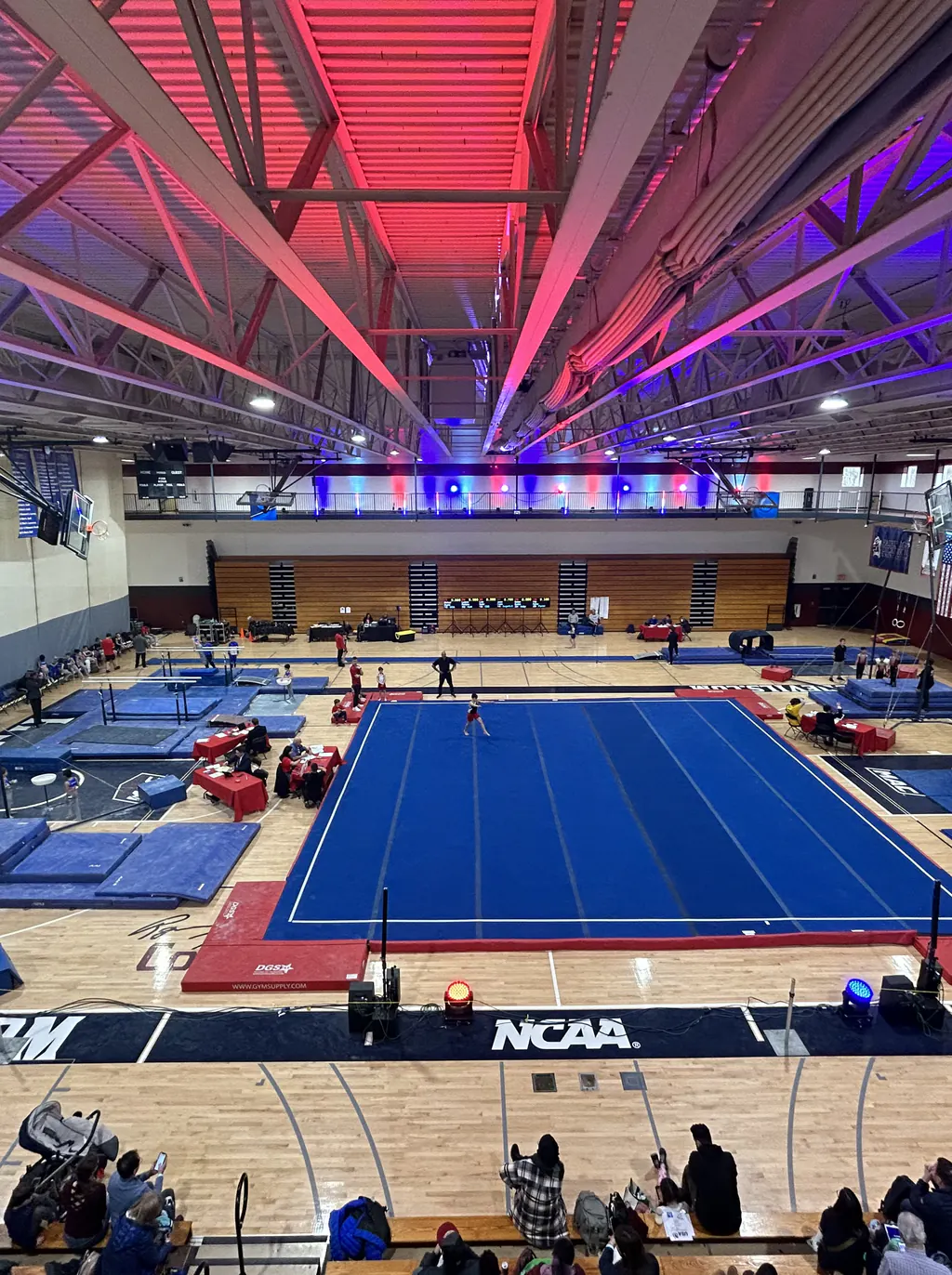 An arena&rsquo;s exposed steel roof washed red and blue from a line of fixtures along the mezzanine rail, above a competition floor and banked seating.