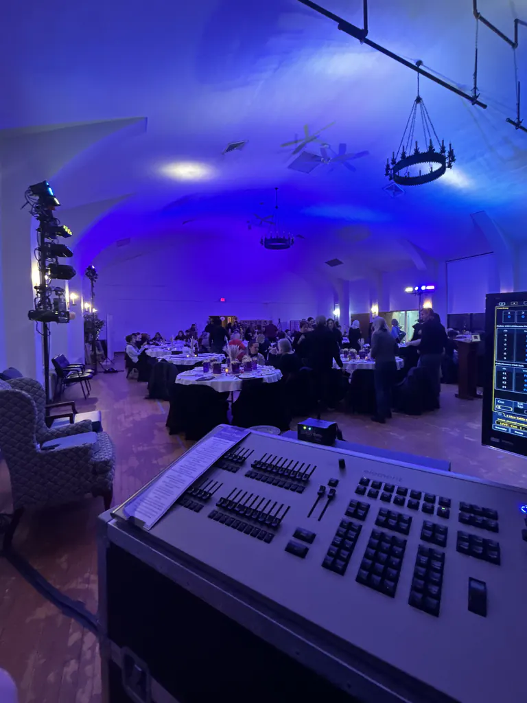 A function room with a vaulted ceiling washed blue, round banquet tables set with linen and centerpieces, guests standing among them, a stand of lighting fixtures at the left, and a lighting console with its monitor in the foreground.